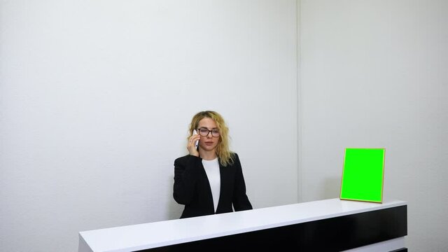 Blonde girl with eyeglasses wearing black jacket standing at reception and talking on phone, mockup screen standing on counter. Concierge communicating with hotel visitors. Concept of hospitality