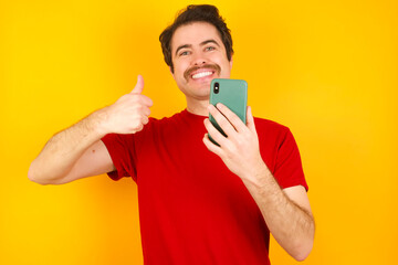 Portrait of Young Caucasian man wearing red t-shirt standing against yellow background using and texting with smartphone  happy with big smile doing ok sign, thumb up with fingers, excellent sign