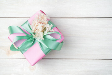 A gift in a pink package with a ribbon decorated with flowers on a white wooden background. Top view, with space to copy. The concept of holiday backgrounds.