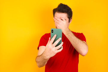 Young Caucasian man wearing red t-shirt standing against yellow background looking at smart phone feeling sad holding hand on face.