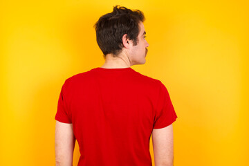 The back side view of a Young Caucasian man wearing red t-shirt standing against yellow background. Studio Shoot.