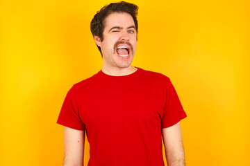 Fototapeta premium Young Caucasian man wearing red t-shirt standing against yellow background winking looking at the camera with sexy expression, cheerful and happy face.