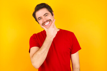 Young Caucasian man wearing red t-shirt standing against yellow background looking confident at the camera smiling with crossed arms and hand raised on chin. Thinking positive.