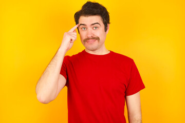 Fototapeta premium Young Caucasian man wearing red t-shirt standing against yellow background tries to memorize something, keeps fore finger on temple, reminds information for exam,