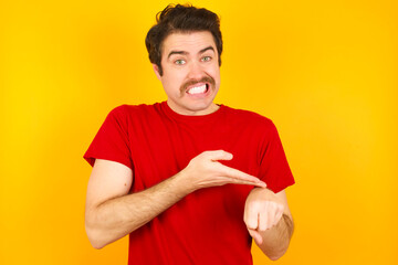 Young Caucasian man wearing red t-shirt standing against yellow background In hurry pointing to...