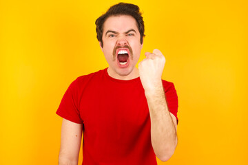 Young Caucasian man wearing red t-shirt standing against yellow wall angry and mad raising fist...