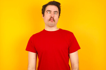 Obraz premium Young Caucasian man wearing red t-shirt standing against yellow wall making fish face with lips, crazy and comical gesture. Funny expression.