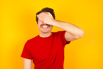 Fototapeta na wymiar Young Caucasian man wearing red t-shirt standing against yellow wall smiling and laughing with hand on face covering eyes for surprise. Blind concept.