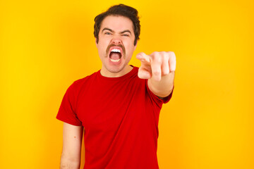 Young Caucasian man wearing red t-shirt standing against yellow wall pointing displeased and frustrated to the camera, angry and furious ready to fight with you.