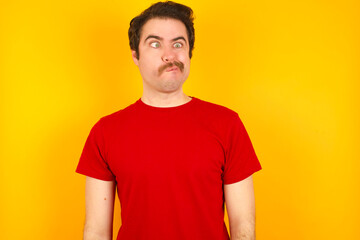 Fototapeta premium Young Caucasian man wearing red t-shirt standing against yellow wall making grimace and crazy face, screaming out of control, funny lunatic expressing freedom and wild.