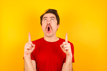 Young Caucasian man wearing red t-shirt standing against yellow wall amazed and surprised looking up and pointing with fingers and raised arms.