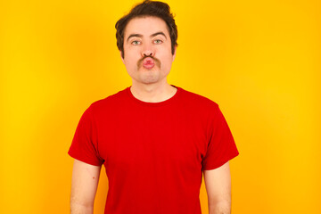 Obraz premium Shot of pleasant looking Young Caucasian man wearing red t-shirt standing against yellow wall, pouts lips, looks at camera, Human facial expressions