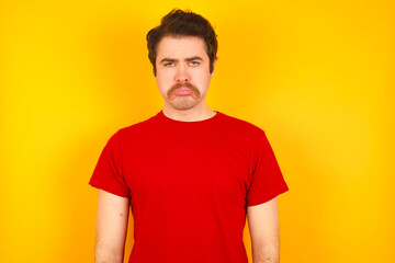 Fototapeta premium Displeased upset Young Caucasian man wearing red t-shirt standing against yellow wall frowns face as going to cry, being discontent and unhappy as can't achieve goals, Disappointed model has troubles