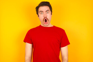 Obraz premium Oh my God. Surprised Young Caucasian man wearing red t-shirt standing against yellow wall stares at camera with shocked expression exclaims with unexpectedness,