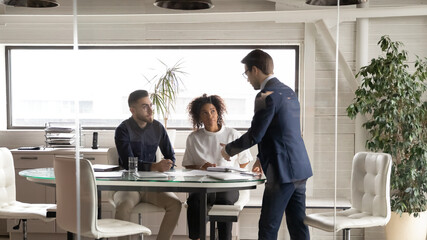 Diverse employees listening to team leader at meeting in boardroom behind glass wall, confident executive training staff, giving instructions, explaining strategy, business partners brainstorming