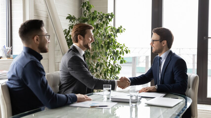 Happy business partners shaking hands, greeting or making successful great commercial deal after successful negotiations, smiling diverse employees sitting at table in modern boardroom, acquaintance