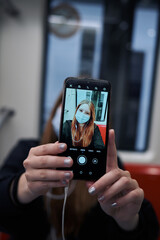 A beautiful white Latina woman using the city subway using her mobile phone to take a selfie photo with her cell phone wearing a mask and headphones
