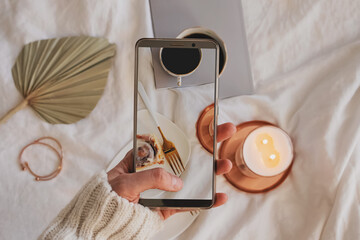 Woman hand taking picture of composition with cinnamon bun, coffee and burning candle