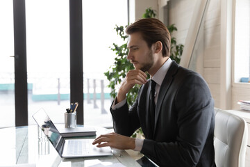 Side view thoughtful businessman looking at laptop screen, pondering task, online project strategy or financial report, pensive entrepreneur touching chin, using computer, sitting at desk in office