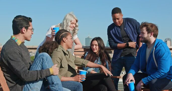 Group of young co-workers hanging out on rooftop patio laughing and having a drink, sharing images on mobile devices 