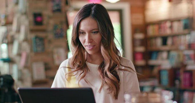 Portrait of young woman sitting at counter of coffee shop bookstore using laptop computer, then looking up into camera smiling.