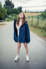 Obraz premium Young woman with eyes closed towards the sky, wearing a blue raincoat and summer clothes standing in the rain