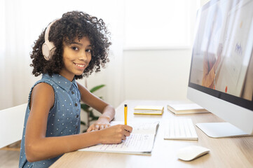 Cute African school girl enjoying e-learning, looks at the camera with a cheerful smile watching online classes, tutorial on trendy PC at home, using interesting app for studying on the distance