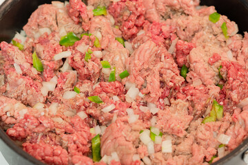 Raw ground meats sitting on a bed of raw onions and green bell peppers in non-stick skillet on a stove top in a domestic kitchen.