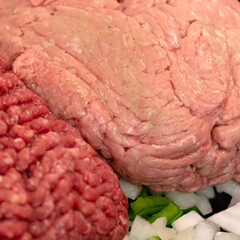 Raw ground meats sitting on a bed of raw onions and green bell peppers in non-stick skillet on a stove top in a domestic kitchen.