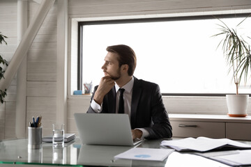 Thoughtful businessman touching chin, looking to aside, pondering difficult task, sitting at desk with laptop in office, pensive executive manager working on online project, developing strategy