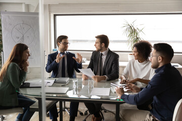 Diverse employees listening to colleague at meeting, sitting at table in boardroom, confident businessman sharing ideas with coworkers at briefing, business partners discussing project strategy