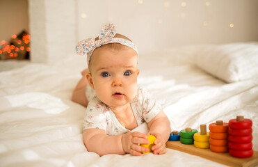 Fototapeta na wymiar a beautiful baby girl in a colored bodysuit and a headband lies on the bed with an educational toy on her head and looks at the camera
