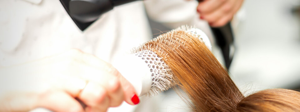 Close up of hairdresser drying long red hair with a hairdryer and round brush in a beauty salon