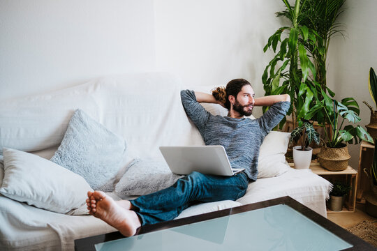 Man with hands behind head sitting with laptop on sofa at home