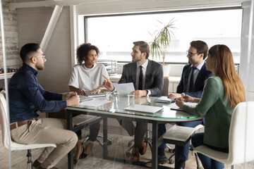 Interested diverse colleagues listening to leader, discussing statistics, sitting at table in modern boardroom, mentor coach explaining strategy, giving instructions to workers at briefing