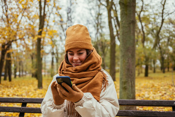 Smiling young woman using mobile phone while sitting on bench at autumn park