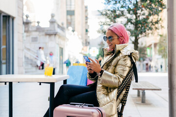 Smiling pink hair woman using mobile phone while sitting at sidewalk cafe