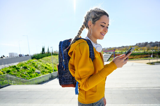 Smiling athlete with backpack using mobile phone while standing on footpath