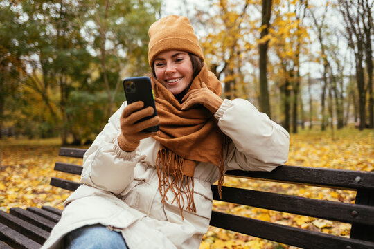 Happy young woman with scarf around neck using smart phone on bench at autumn park