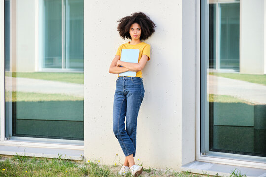 Female student holding book while leaning on wall at university campus