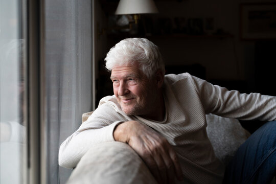 Smiling senior man looking through window while sitting on sofa at home