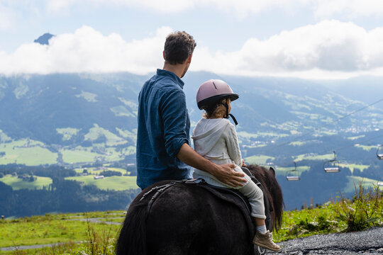 Father walking beside little daughter riding pony against mountain valley