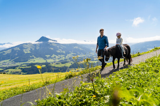Father walking beside little daughter riding pony along alpine footpath