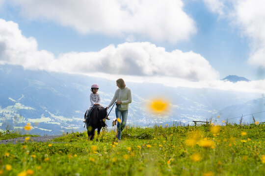 Mother walking beside little daughter riding pony in alpine meadow