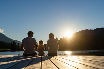 Family with little daughter sitting together at end of lakeshore jetty at sunset