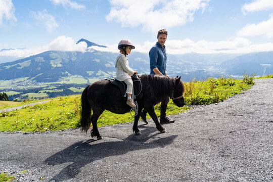 Father walking beside little daughter riding pony against mountain valley