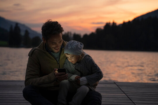 Father and little daughter playing with smart phone on lakeshore jetty at dusk