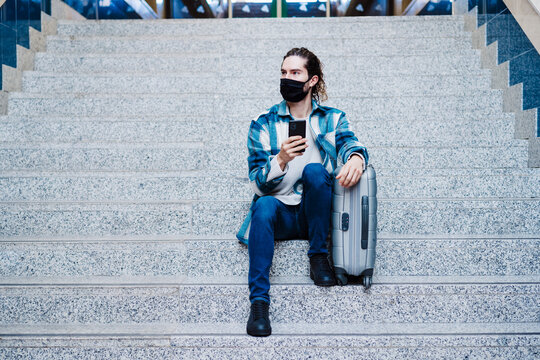 Man wearing protective face mask using mobile phone while sitting with luggage on steps