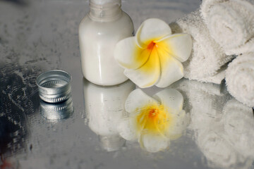white towel body lotion and plumeria flower are reflected in the mirror surface of the wet table