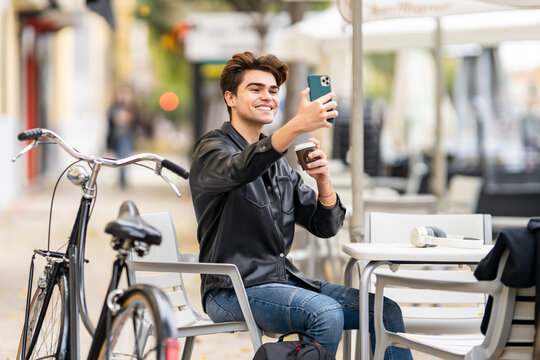 Smiling handsome man taking selfie with coffee cup through smart phone while sitting at sidewalk cafe in city